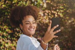 Smiling Black woman taking selfie on phone against trees