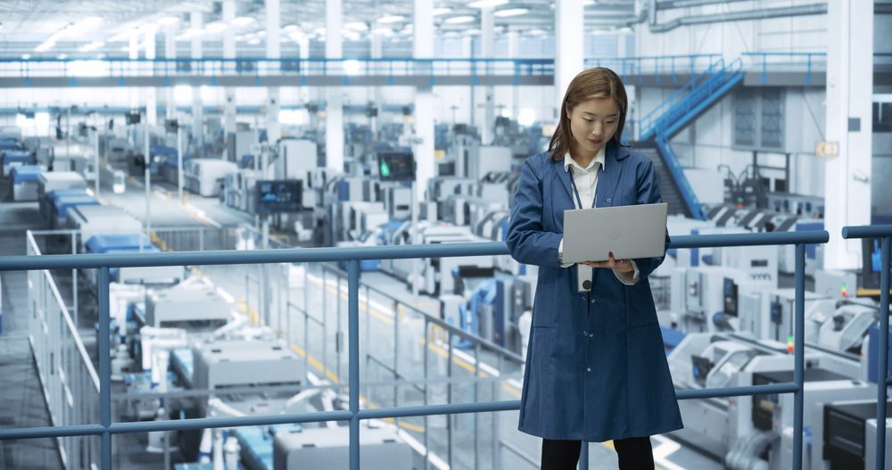 Asian Female Engineer Standing in a Factory Facility, Working on Laptop Computer, Researching and Developing Technologies to Produce Electronics at a Modern Automated Manufacture with Robotic Arms