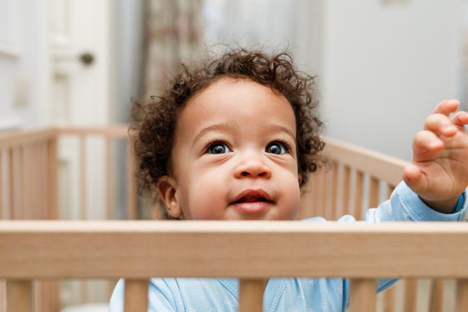 Cute and curious baby boy looking over the top of his crib railing