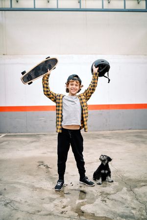Cheerful child raising skateboard with helmet near dog on street