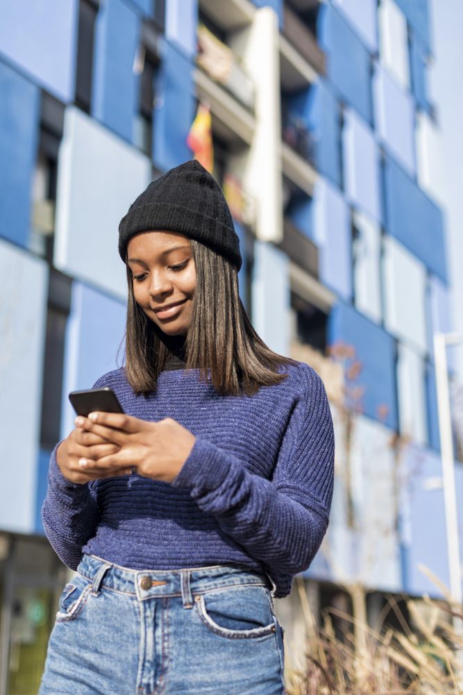 Female in hat and blue sweater checking cell phone outside blue building, vertical