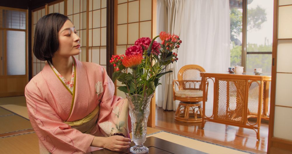 Japanese Female in a Pink Kimono Sitting on Her Knees on Tatami Flooring, Admiring a Vase with a Colorful Flowers Bouquet.