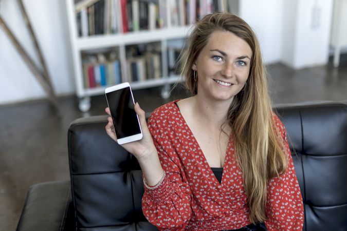 Young blonde woman sitting on sofa and holding mobile phone while smiling at camera