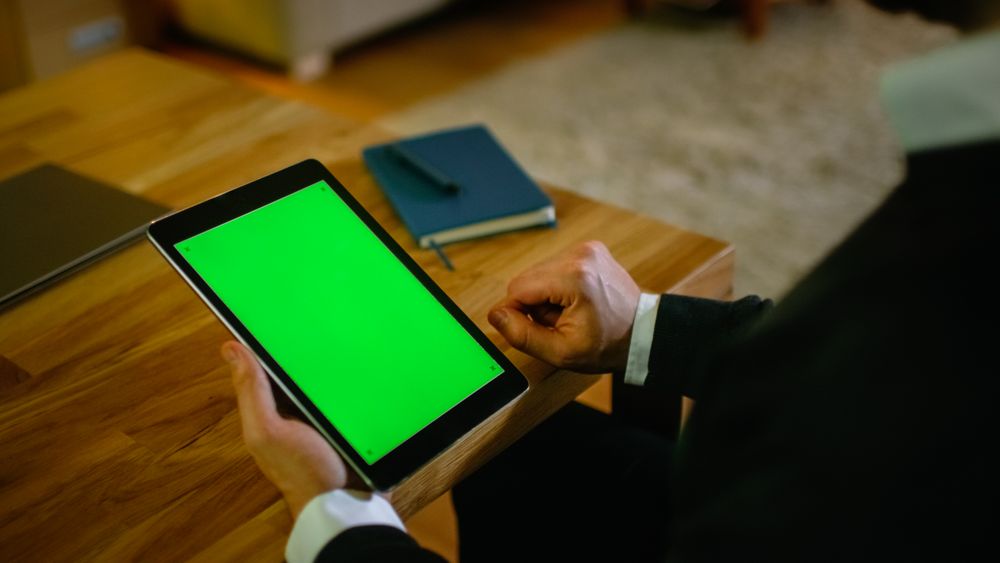 At Home Man Sits at His Desk and Holds Tablet Computer with Green Screen on it.
