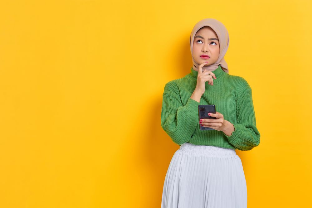 Woman in headscarf holding smart phone looking up while thinking about something