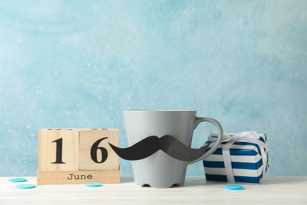 Cup with decorative mustache, gift box and wooden calendar on table against blue background, space for text