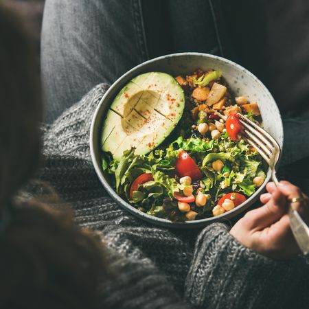 Woman in jeans and sweater eating bowl of fresh salad, top view, square crop