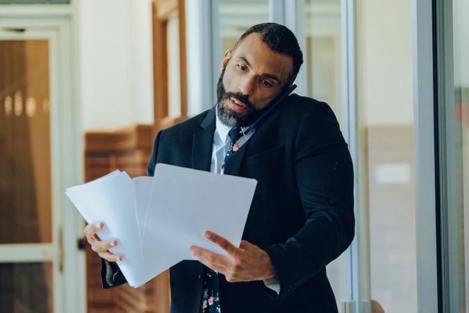 Serious businessman in suit and tie reviewing papers standing in office while on phone