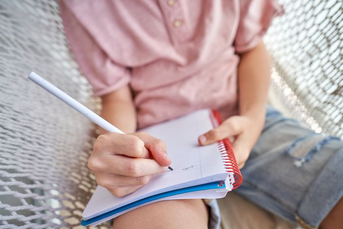 Crop boy lying in hammock and taking notes in notepad