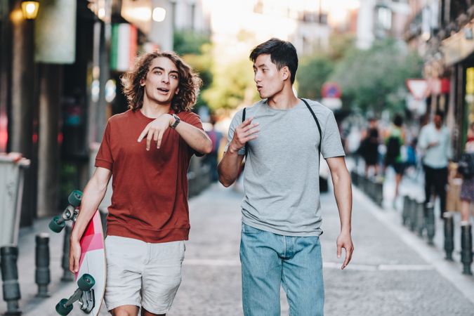 Cool multiracial male friends chatting while walking on street with longboard