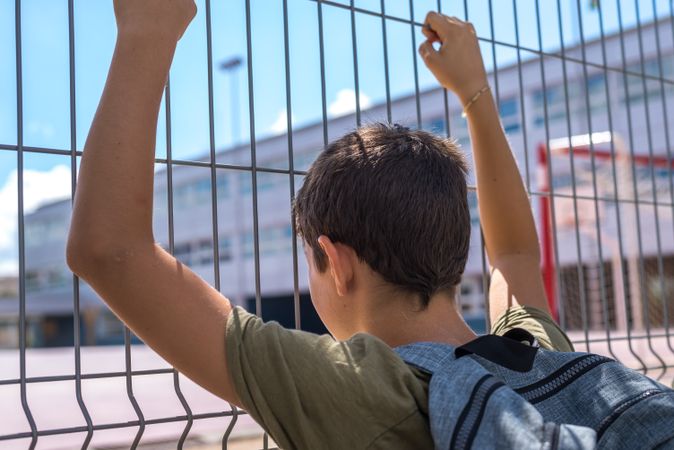 Back of boy holding gate with both hands looking into school yard