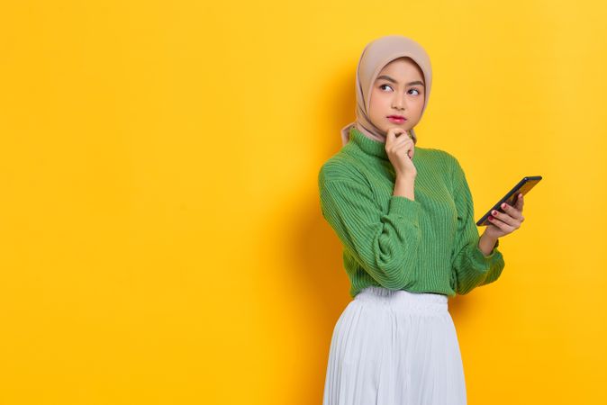 Woman in headscarf looking away from smart phone and thinking