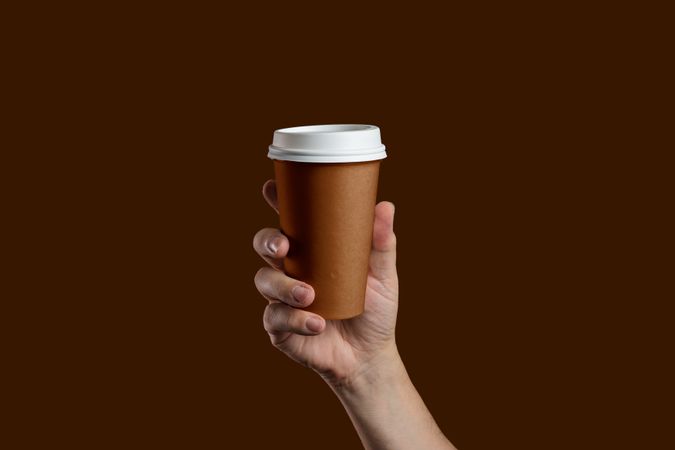 Hand holding a takeaway paper coffee cup on a solid brown background.