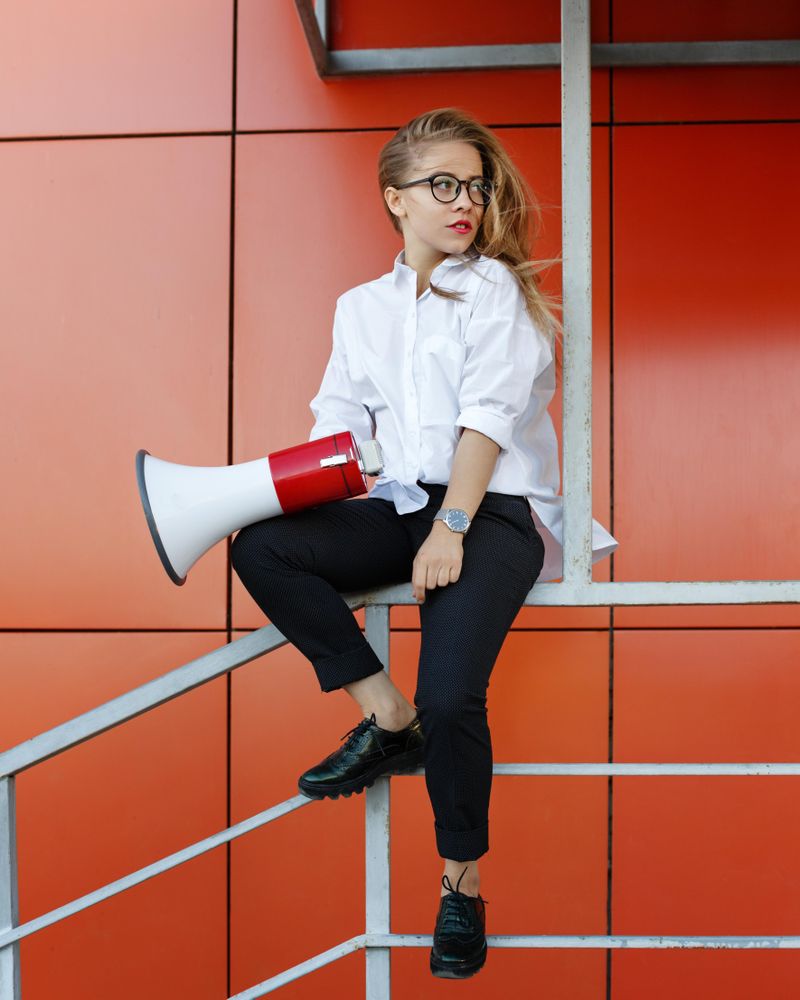Woman in glasses sitting in front of orange wall with megaphone