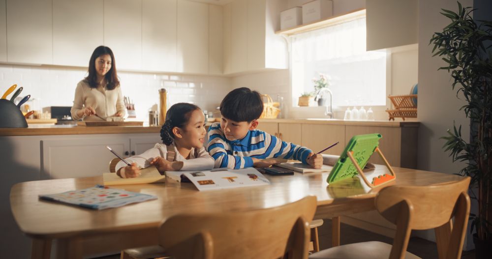 Portrait of a Little Korean Kids Sitting at a Kitchen Table at Home in the Morning While Their Mother is Cooking in the Background.