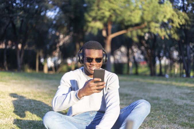 Black male with sunglasses sitting outdoors checking his phone in park