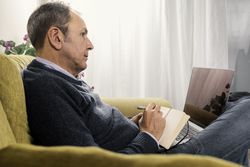 Man in an online therapy session taking notes at home.