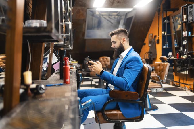 Man in sharp blue suit checking phone in barber’s chair in salon