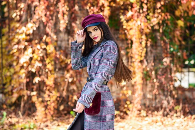 Female in warm winter clothes standing in park with colorful autumn leaves holding her hat