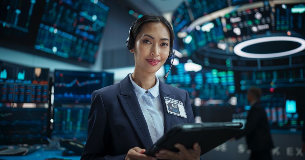 Portrait of a Asian Female Working in an International Stock Exchange Hall: Specialist Wearing Headphones and Using Tablet Computer, Successful Trader Looking at Camera and Smiling