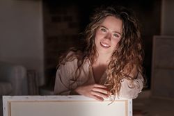 Smiling woman with long curly hair sitting near canvas