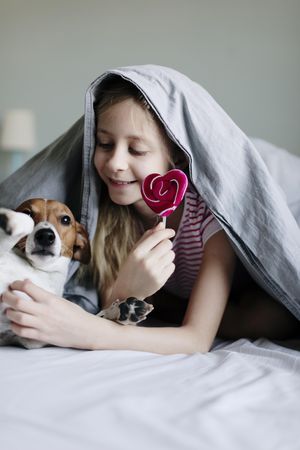 Girl with blanket draped on her head petting her dog while holding a lollipop
