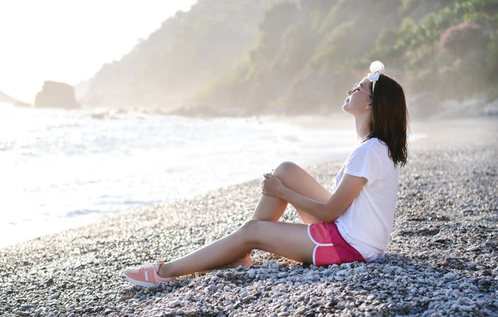 Young woman sitting on pebble beach, eyes closed with face to the sky