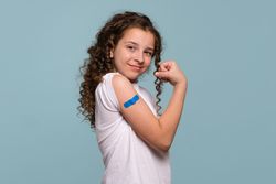 Girl flexing arm with blue bandage after receiving vaccine shot.