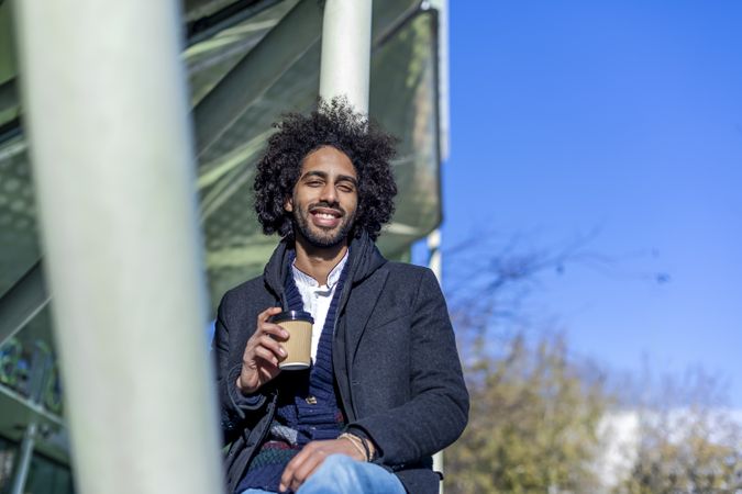 Smiling Black man in jacket with warm beverage sitting balancing between poles outside
