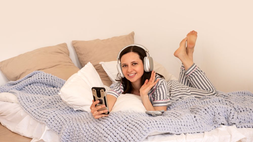 Woman in bed with headphones, smiling and waving.
