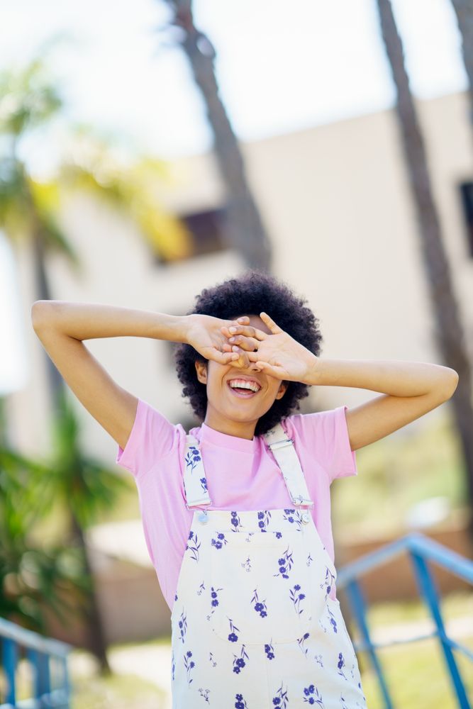 Black woman in floral coveralls covering her eyes with open palms