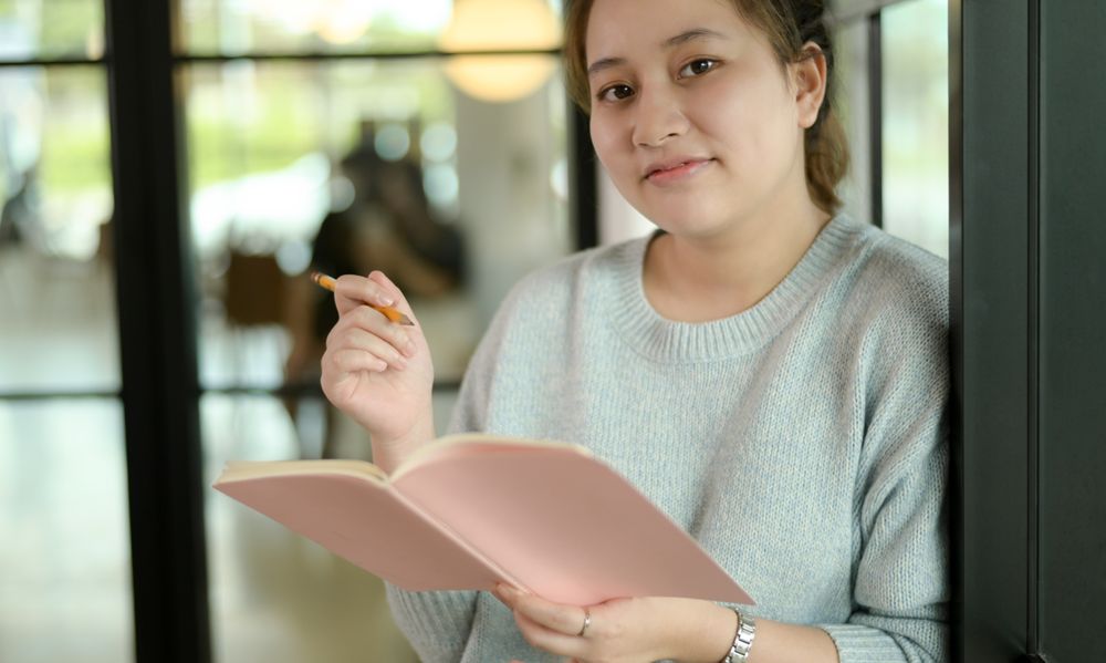 Young woman holding a notebook and pencil leaning against a glass wall.