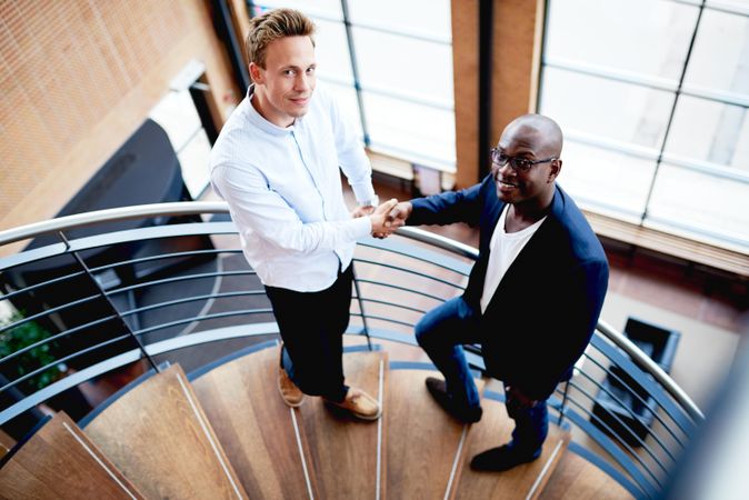 Two smiling businessman shaking hands on staircase and looking at camera