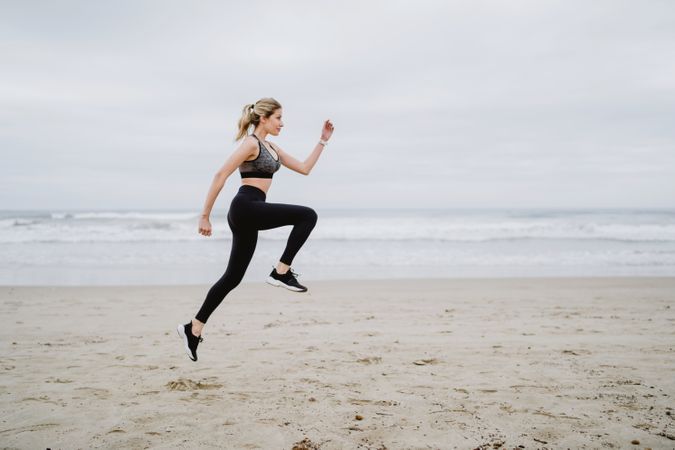 Young female athlete in active wear jogging while jumping at sea beach