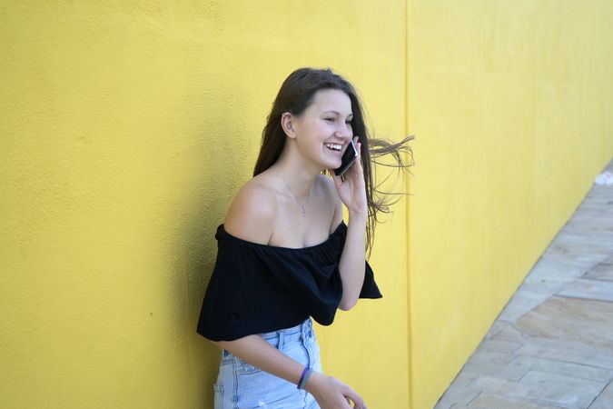 Happy woman leaning on yellow wall speaking on cell phone