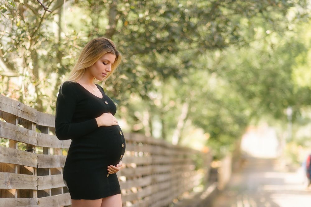 Pregnant woman looking away and touching belly while standing on road near garden in sunny day