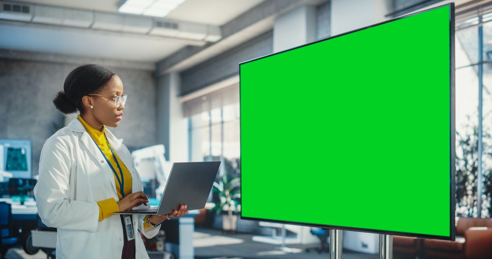 Female Specialist Using a Laptop Computer and Looking at a Green Mock Up Displayed on Big Digital Screen.