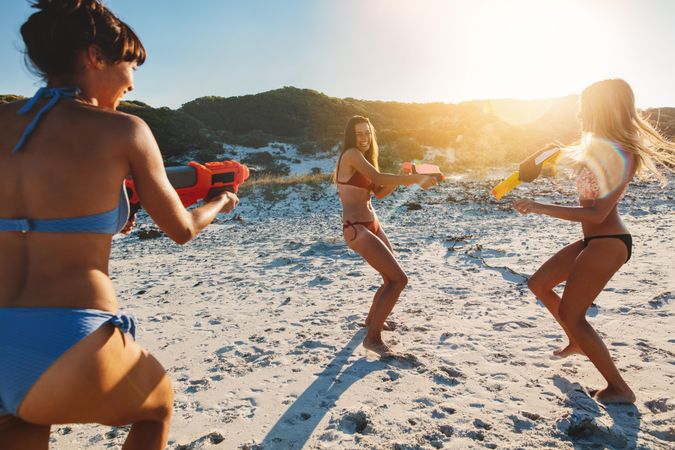 Slender females playing with toy water guns on sandy beach