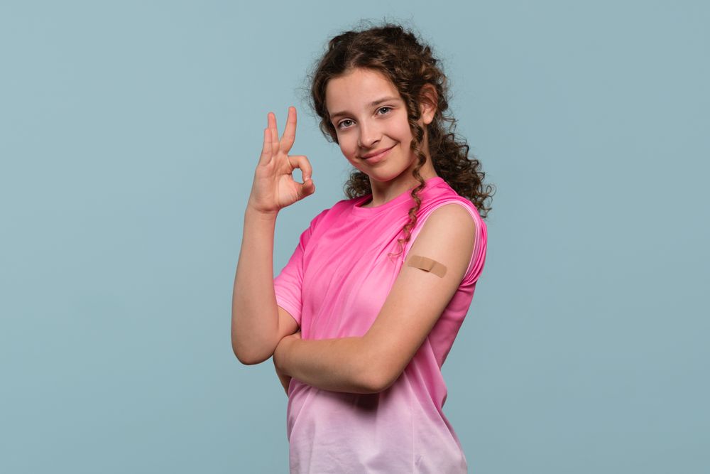 Smiling girl giving "OK" gesture, showing bandage on arm.
