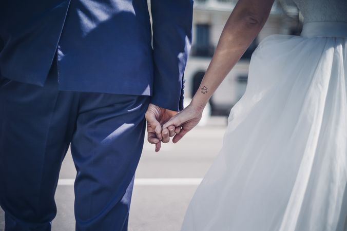 crop man holding on hands woman in wedding dress on street in sunny day