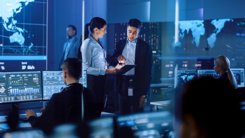 Professionals Collaborate in High-tech Control Room. Two Discuss Data on Tablet, Surrounded by Analysts Monitoring Global Information on Multiple Screens. World Map Display Dominates Background.