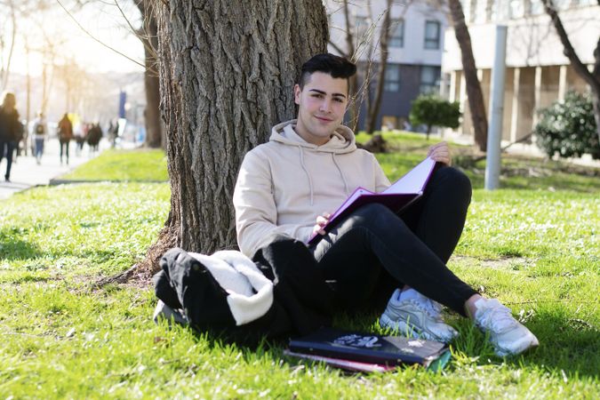 Young smiling man leaning on a tree while reading a book