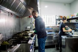 Side view of bearded male chef and woman cooking food over pan and washing dishes in modern kitchen of restaurant