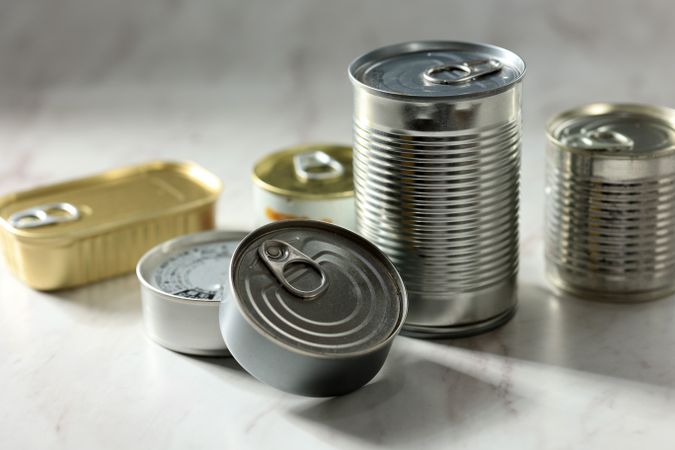 Various Closed Tin Cans with Food Preserves on a Light Gray Background.