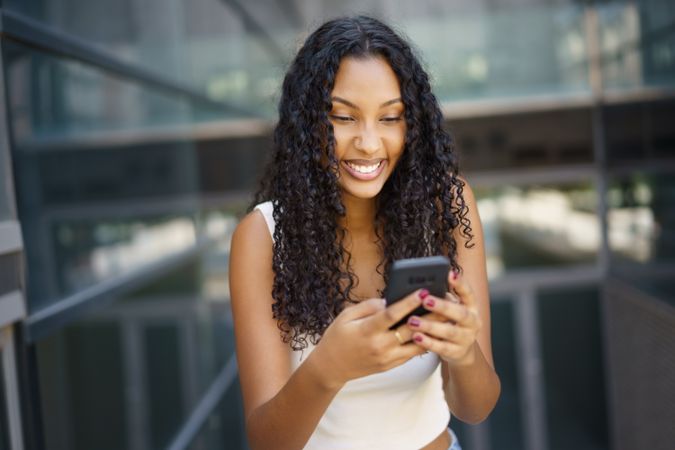 A Young Woman Happily Smiling While Using Her Smartphone Outdoors in Bright Daylight