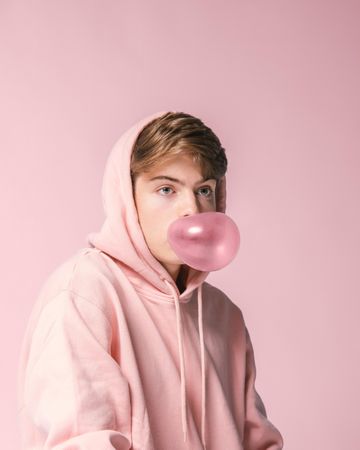 Teenage boy in a pink hoodie blowing bubble gum against pink background