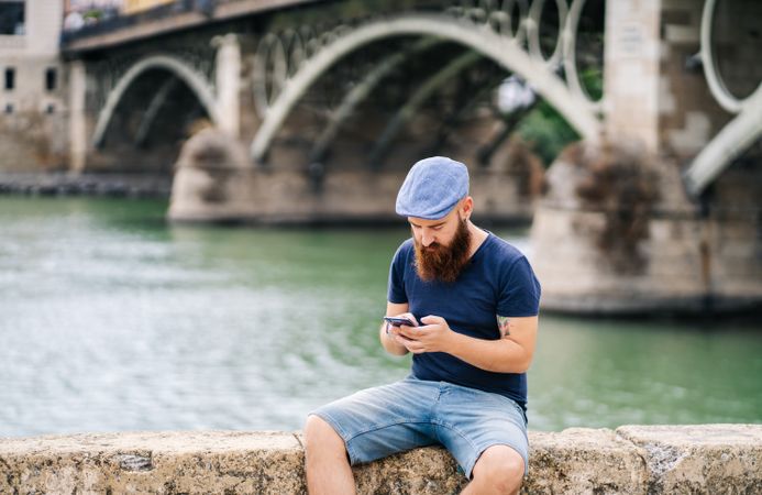 Bearded young man in casual wear using smartphone while walking on quay