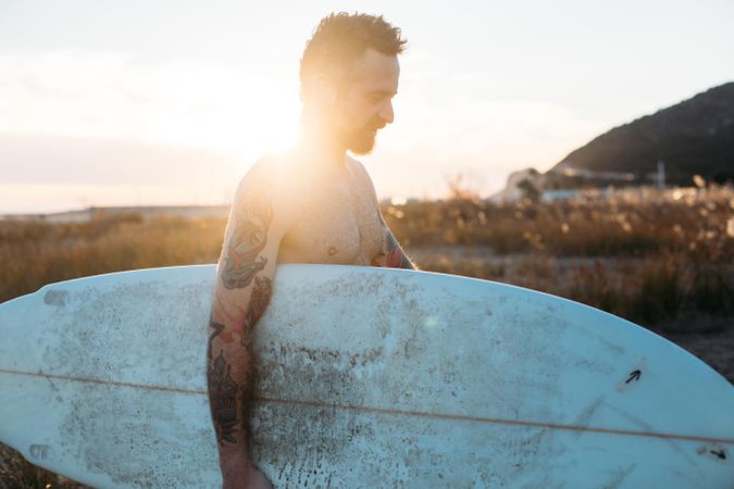 Tattooed man looking down with surfboard under his arm with sun behind him