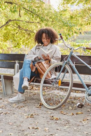 Cheerful Black woman reading book on bench near bicycle in park