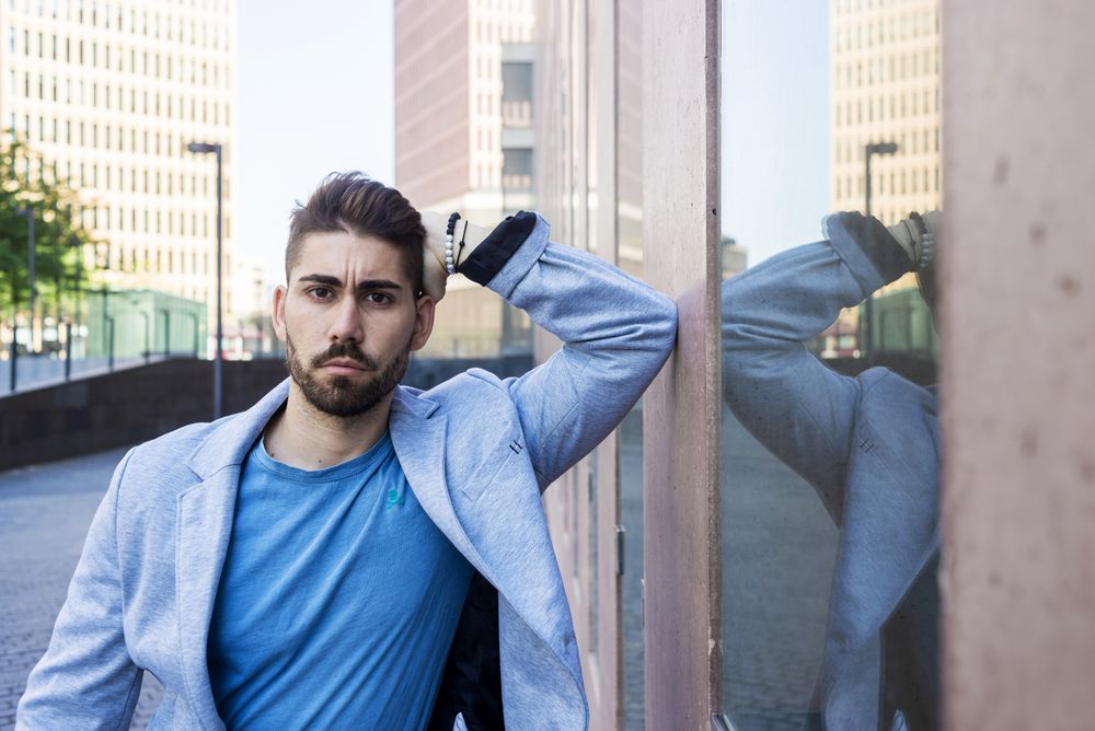 Young man in blazer leaning elbow on a building wall outside looking at camera